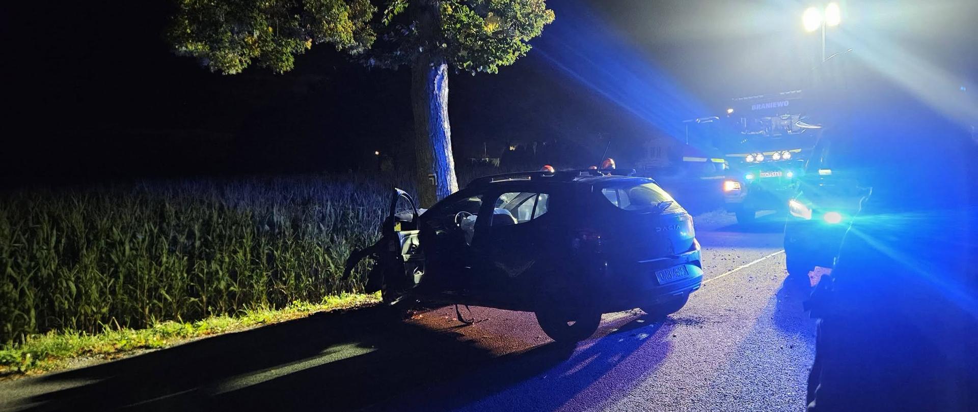 Na zdjęciu widać samochód osobowy po wypadku, stojący na drodze nocą. Auto jest mocno uszkodzone z lewej strony i znajduje się blisko drzewa. W tle widać samochód straży pożarnej z Braniewa oraz intensywne niebieskie światła służb.
