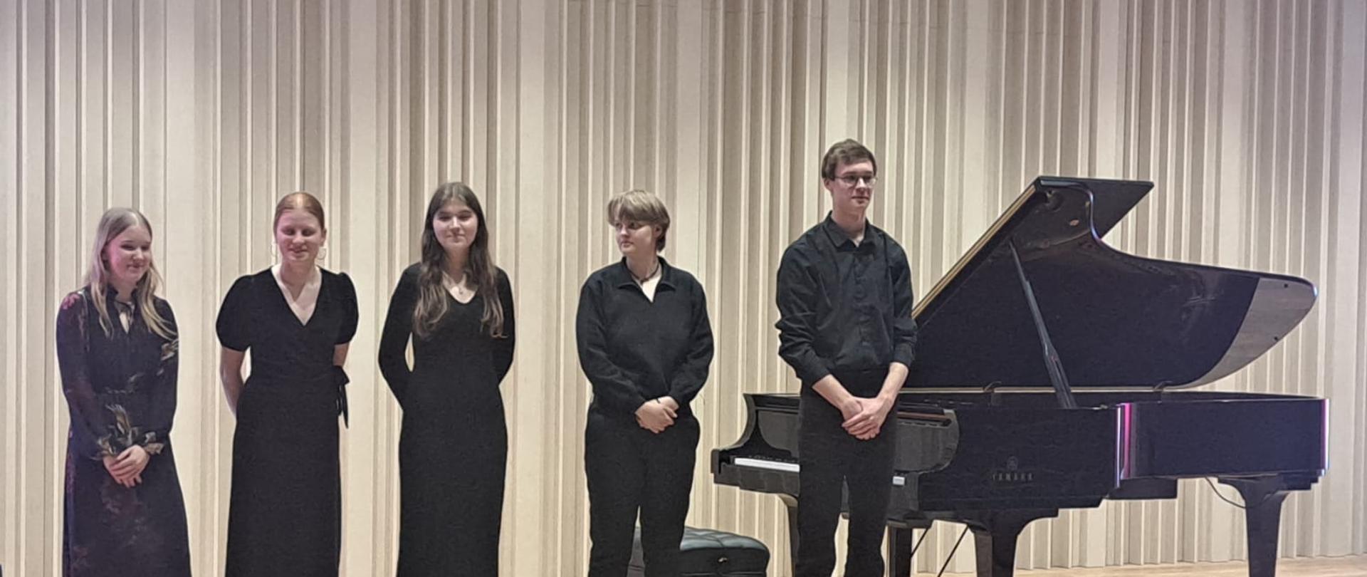 Na zdjęciu pięciu młodych artystów. Wszyscy ubrani na czarno. Po prawej stronie zdjęcia czarny fortepian. Tło obrazka w odcieniach beżu.