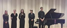 Na zdjęciu pięciu młodych artystów. Wszyscy ubrani na czarno. Po prawej stronie zdjęcia czarny fortepian. Tło obrazka w odcieniach beżu.