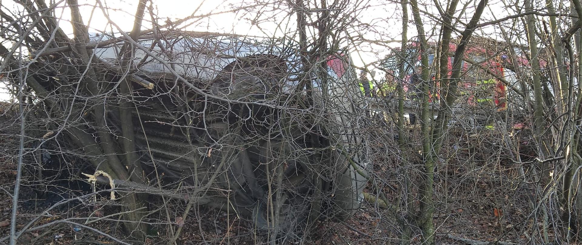 samochód osobowy koloru srebrnego opat o drzewa i krzaki na skarpie wiaduktu kolejowego za nim widoczny w tle czerwony samochód straży pożarnej oraz strażacy.