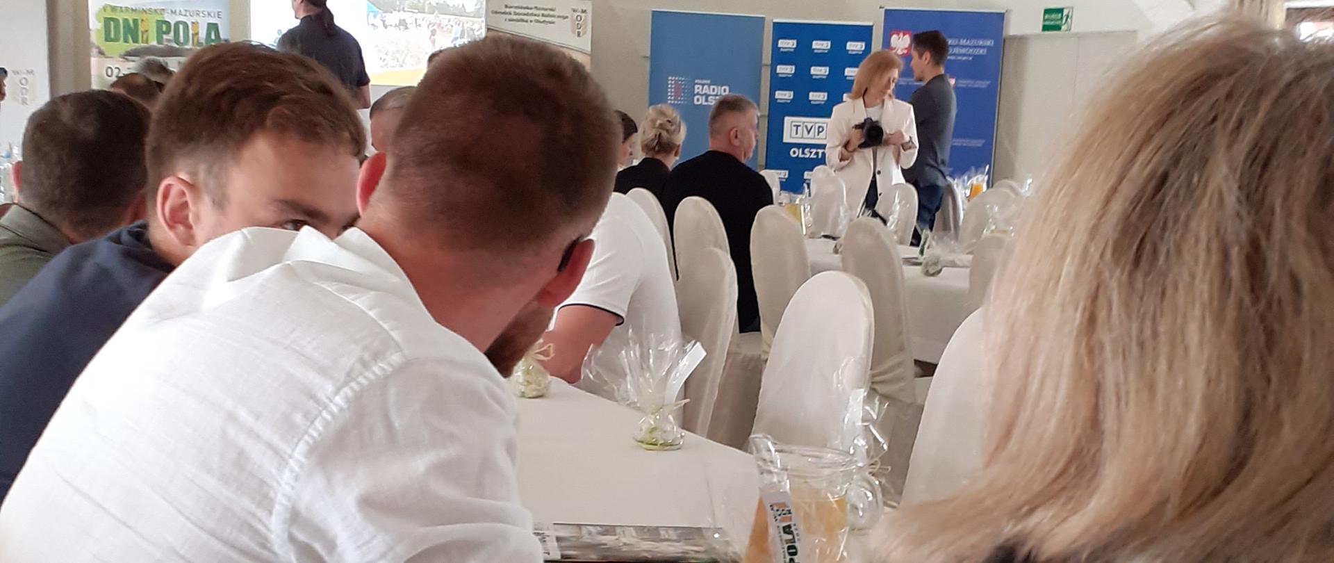 Wnętrze jasnej Sali konferencyjnej z białym sufitem i widocznymi belkami
konstrukcyjnymi. Na środku sali wisi czarny, ozdobny żyrandol z kutego metalu. Na pierwszym planie
tyłem widoczna kobieta z długimi blond włosami w ciemnej bluzie z napisem „Państwowa Inspekcja
Ochrony Roślin i Nasiennictwa”. Obok niej siedzi mężczyzna w białej koszuli i niebieskich dżinsach. W
tle widać inne osoby siedzące przy stołach nakrytych białymi obrusami. Na stołach znajdują się
szklanki z napojami i dekoracje. Z przodu sali osoba prowadząca prezentację stoi obok ekranu i kilku
kolorowych banerów informacyjnych. 