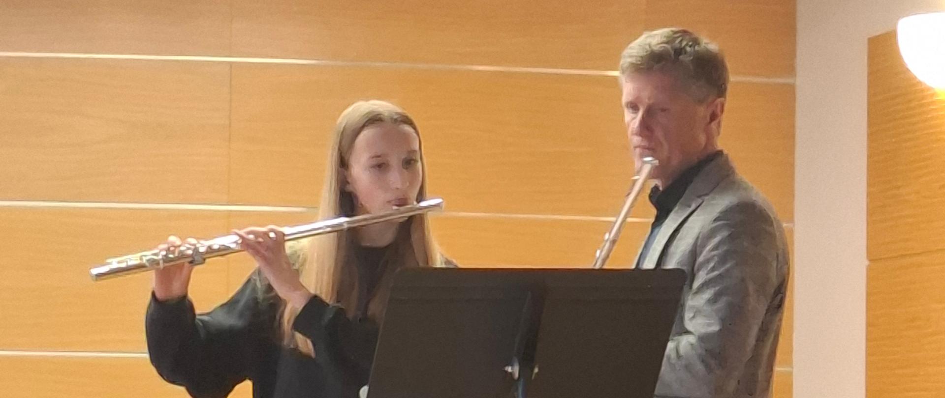 Zdjęcie przedstawia dwoje flecistów podczas wspólnej lekcji w sali kameralnej Po lewej stronie stoi uczennica w czarnym stroju, a po prawej prowadzący warsztaty w szarej marynarce i jasnych spodniach. Oboje grają na poprzecznych fletach srebrnego koloru. Pomiędzy muzykami znajduje się wysoki pulpit na nuty. W tle widać fragment czarnego fortepianu oraz ściany wyłożone jasnymi, drewnianymi panelami. Zdjęcie zostało wykonane z poziomu widowni, co sugerują widoczne na dole oparcia krzeseł. Całość emanuje atmosferą skupienia i muzycznej współpracy.