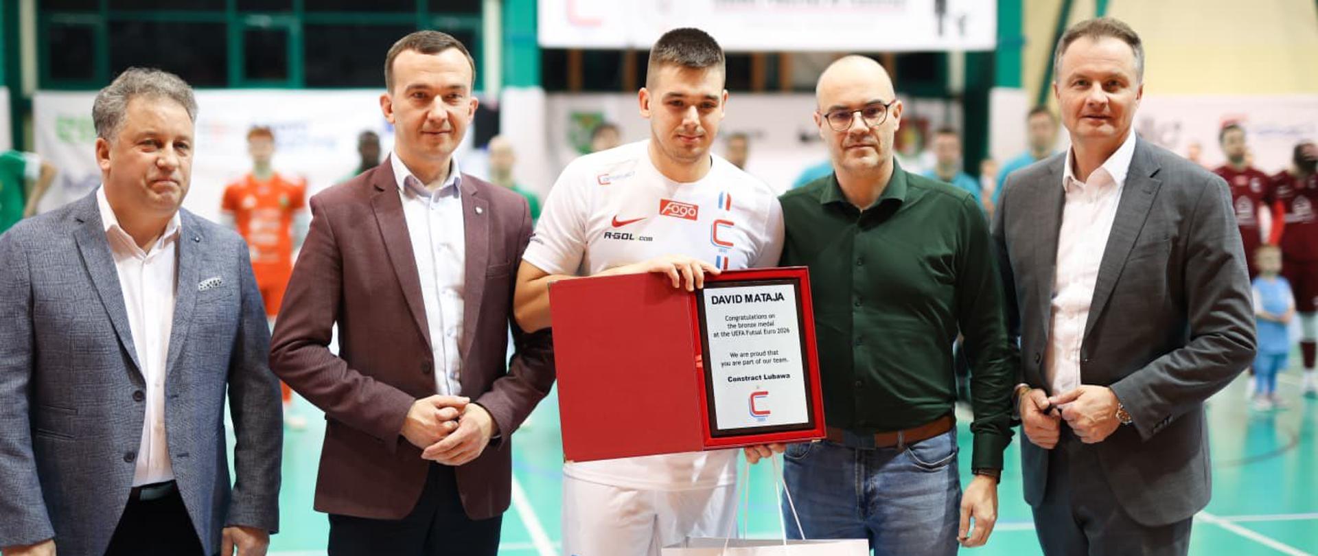 Na boisku grupa mężczyzn, w tym wicewojewoda trzyma certyfikat, wyrażając radość z osiągnięcia.