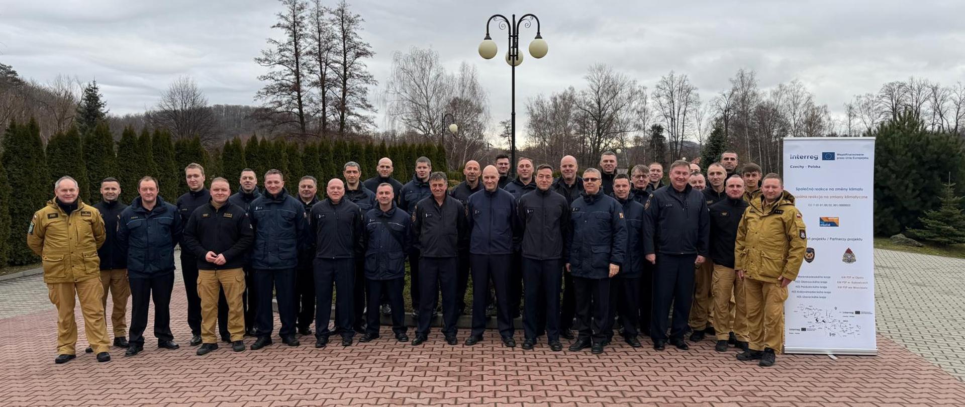 Uczestnicy stażu wymiennego z PSP i HZS pozują do wspólnego zdjęcia obok roll‑upu projektu „Wspólna reakcja na zmiany klimatu”, realizowanego w ramach współpracy transgranicznej Czech i Polski.
