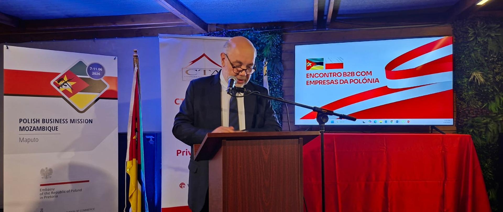 Chargé d’Affaires Jacek Chodorowicz speaking at the Global Gateway Forum 2026 in Maputo; in the background are two roll-up banners, including one from the Polish Chamber of Commerce; the inscription on the second banner is not visible; in the center is the Mozambican flag; to the right is a screen displaying the words: Encontro B2B com Empresas da Polonia. The screen is placed on a table covered with red cloth.
