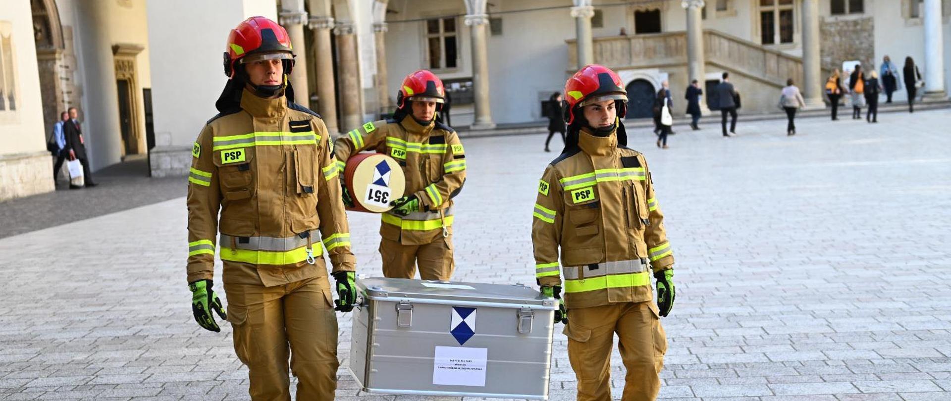 Trzech strażaków PSP w ubraniach specjalnych z hełmami niesie skrzynię transportową i przewód ochronny oznaczony symbolem dóbr kultury, przygotowując się do działań zabezpieczających.