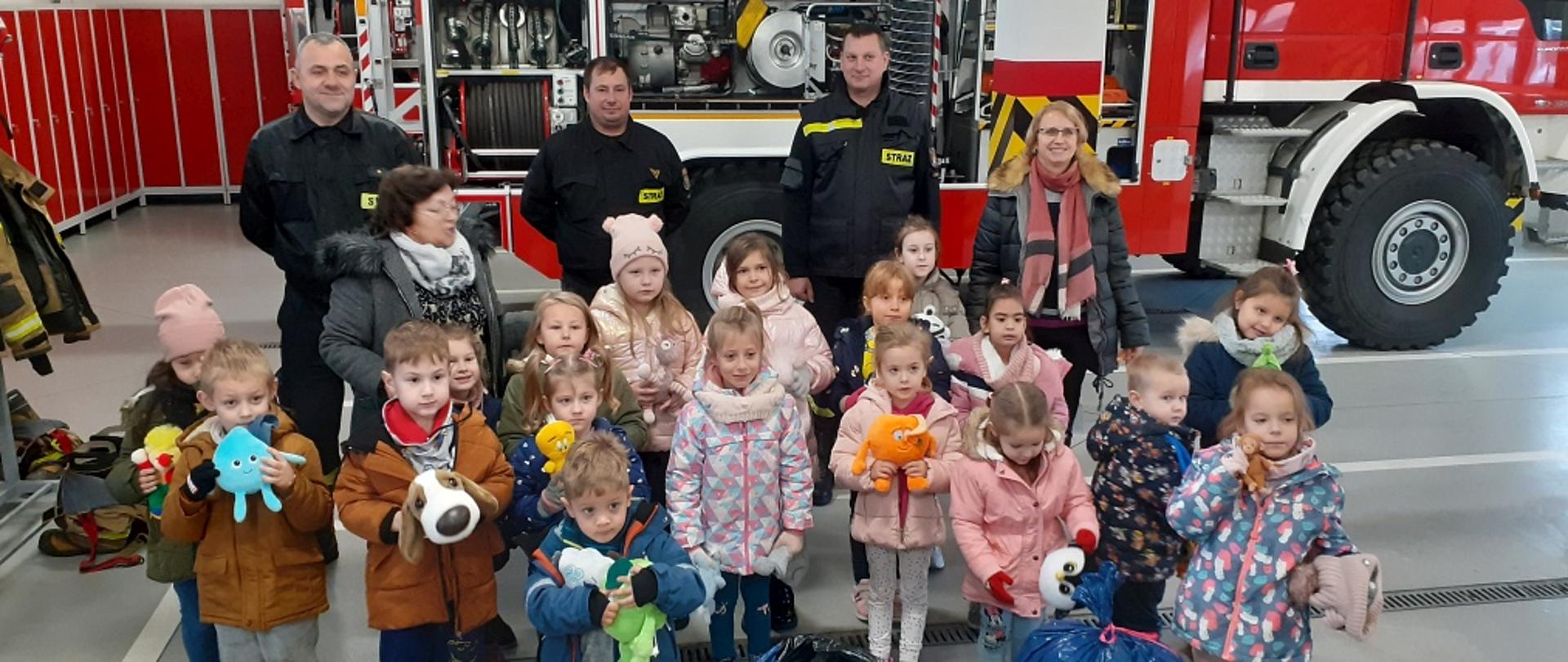 Na zdjęciu widać wycieczkę dzieci z pluszakami w JRG Kolno, w tle samochód pożarniczy.