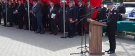 Powiatowe obchody Dnia Strażaka w Suwałkach - przemówienie podlaskiego komendanta wojewódzkiego PSP, w tle zaproszeni goście stojcy pod czerwonym namiotem PSP
