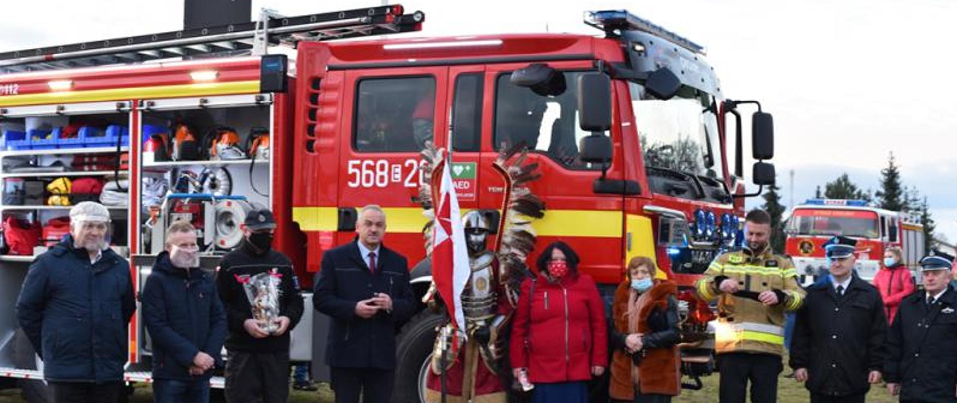 Zdjęcie przedstawia nowy pojazd pożarniczy GBA MAN z Jednostki OSP Chojny oraz przedstawicieli Samorządu Gminnego, Rady Sołeckiej, druhów z jednostek OSP z terenu Gminy Lututów i husarza trzymającego chorągiew.