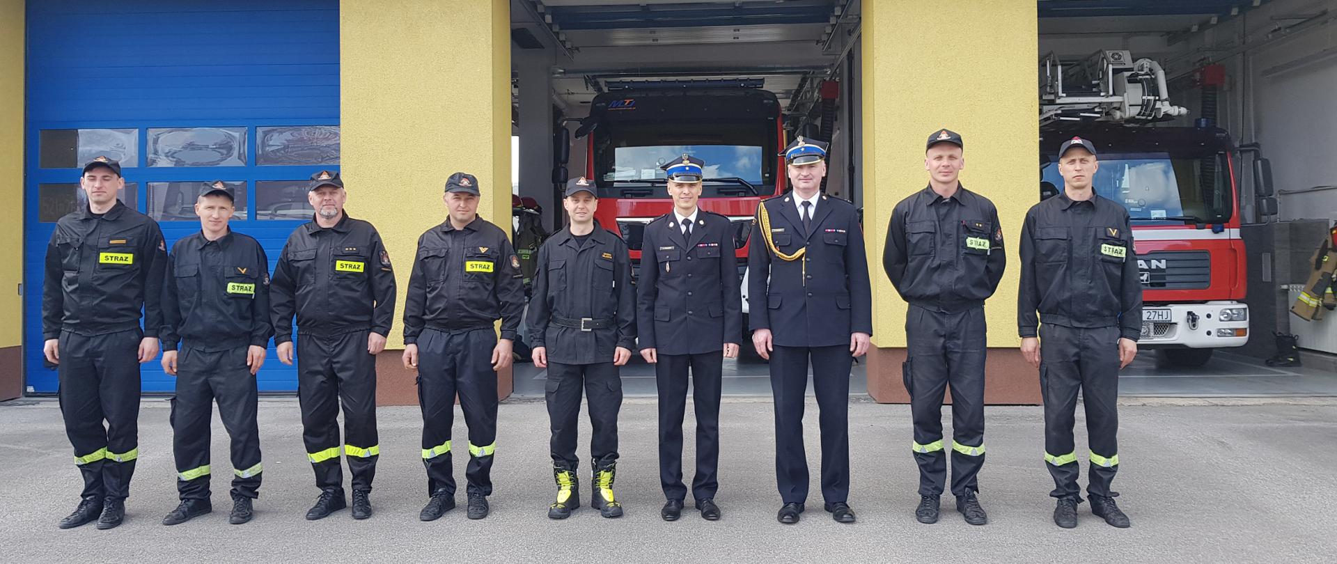 strażacy stoją na tle wozów pożarniczych, w środku stoją w mundurach dwóch strażaków
