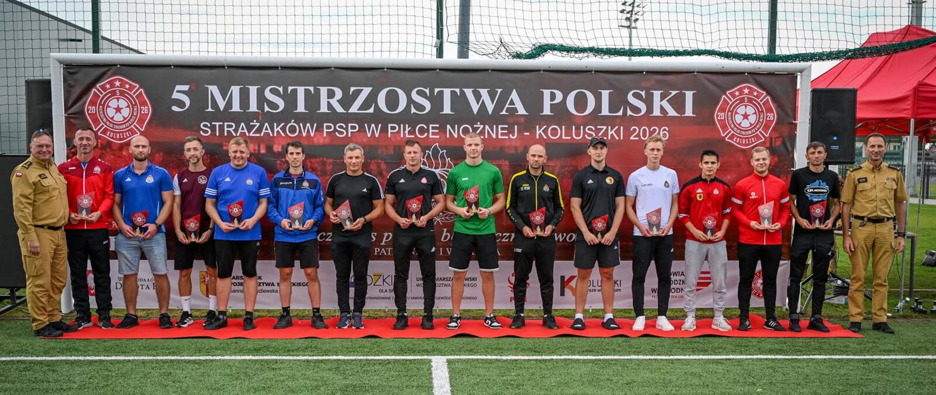 Uczestnicy V Mistrzostw Polski Strażaków PSP w Piłce Nożnej stoją na boisku z pamiątkowymi statuetkami. Po bokach grupy stoją funkcjonariusze PSP. W tle baner mistrzostw w Koluszkach.