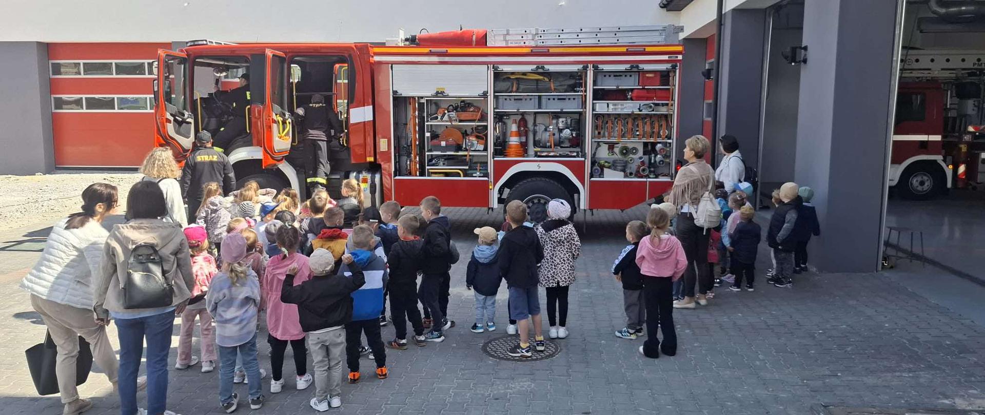 Na zdjęciu widoczna jest duża grupa dzieci, stojąca w rzędach przed czerwoną remizą strażacką. W tle stoi duży czerwony wóz strażacki z otwartymi drzwiami, do którego zaglądają strażacy. Dzieci są pod opieką dorosłych, którzy również stoją obok nich. Całość sugeruje wycieczkę edukacyjną w jednostce straży pożarnej, gdzie dzieci mogą zapoznać się ze sprzętem i pracą strażaków.