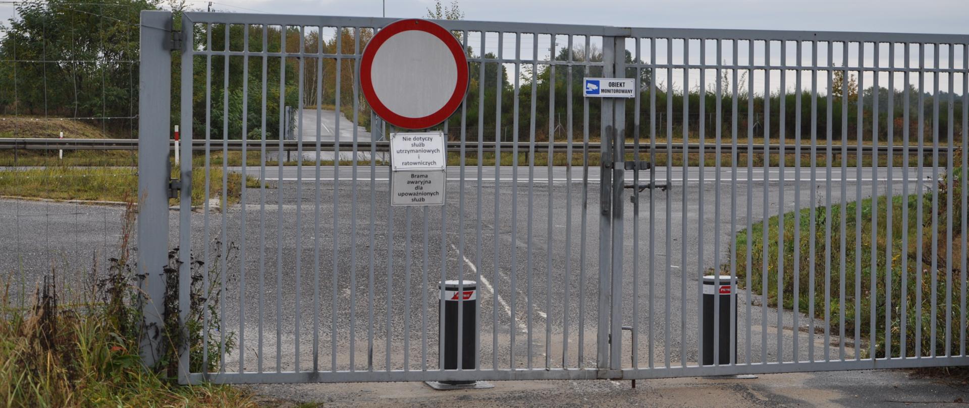 Zamknięta, szara metalowa brama ze znakiem zakazu ruchu oraz informacją, że wjazd dotyczy tylko służb ratowniczych i utrzymaniowych. Na bramie wisi tabliczka „Obiekt monitorowany”, a bezpośrednio za nią stoją dwa słupki blokujące wjazd na pustą drogę. Po drugiej stronie drogi, taka sama brama, ale otwarta.