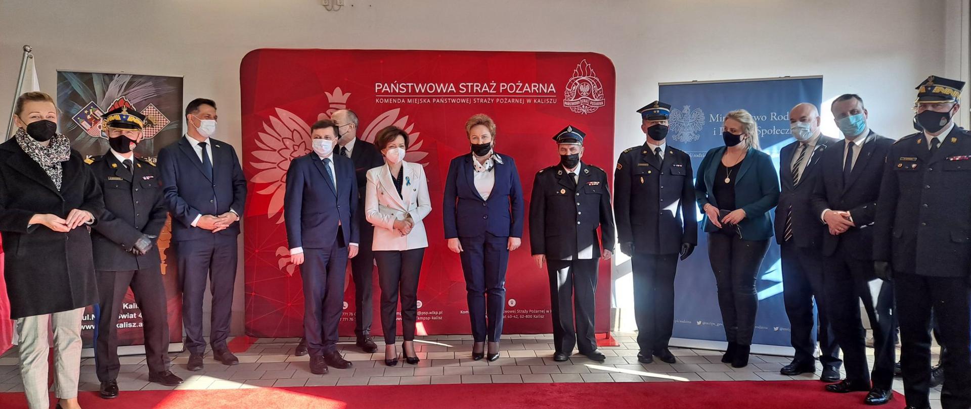 Zdjęcie przedstawia uroczystość wręczenia promes na zakup samochodów gaśniczych, które odbyło się w Komendzie Miejskiej PSP w Kaliszu. Na zdjęciu widać zaproszonych gości: parlamentarzystów, włodarzy gmin i kierownictwo PSP. Zdjęcie wykonane w garażu Komendy Miejskiej PSP w Kaliszu w porze dziennej.