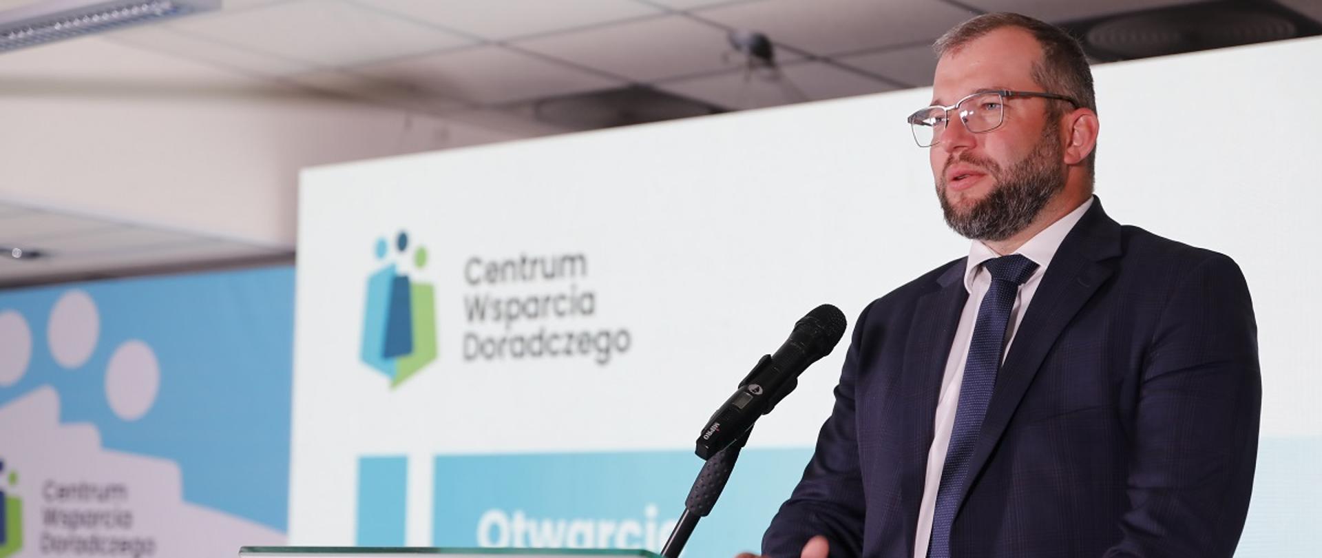 Minister funduszy i polityki regionalnej Grzegorz Puda podczas konferencji podsumowującej efekty działania projektu Centrum Wsparcia Doradczego Plus, minister stoi przy mównicy i przemawia do mikrofonu