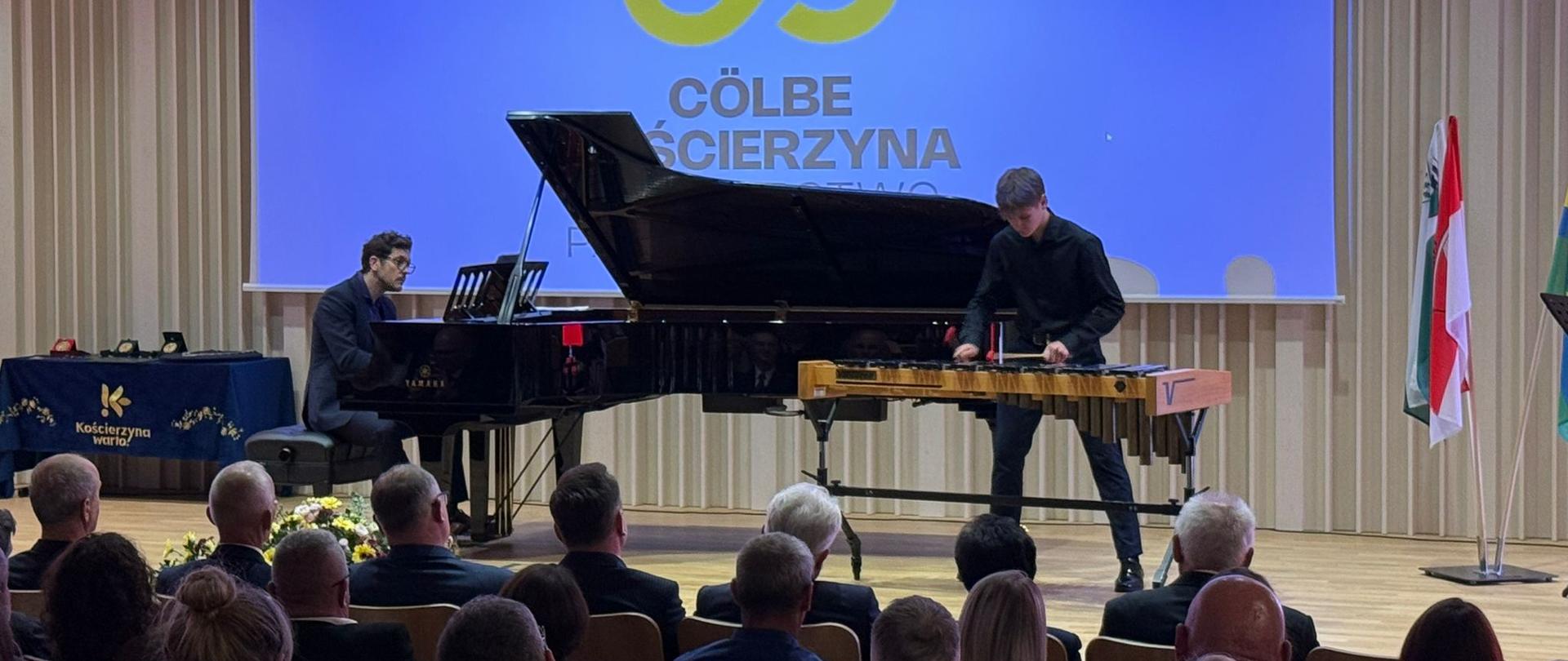 Młody perkusista ubrany na czarno występuje na scenie podczas uroczystości partnerskiej miast Kościerzyna–Cölbe. Gra na wibrafonie, obok niego przy czarnym fortepianie mężczyzna w okularach i czarnym garniturze. W tle widoczny jest duży ekran z logo 35-lecia partnerstwa oraz mównica z mikrofonem. Na widowni siedzą uczestnicy wydarzenia.