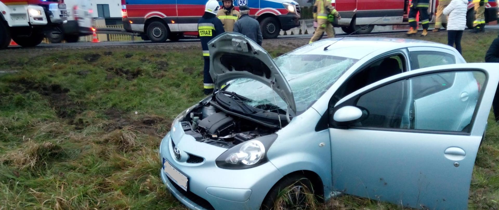 Zdjęcie przedstawia samochód osobowy stojący na przydrożnej łące. W tle, na jezdni widać stojące pojazdy służb ratowniczych: dwa ambulanse i auto pożarnicze
