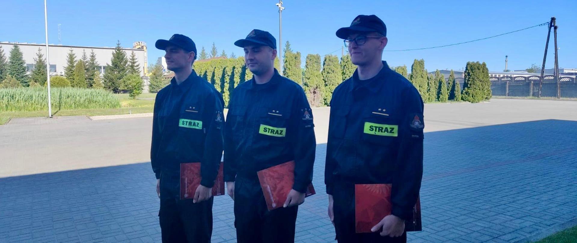 Trzech funkcjonariuszy w umundurowaniu koszarowym stoi na placu. Każdy z nich w lewej ręce trzyma czerwoną teczkę.