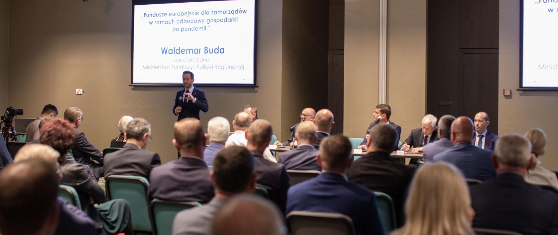 Na tle sali, gdzie siedzą ludzie, stoi z mikrofonem Waldemar Buda