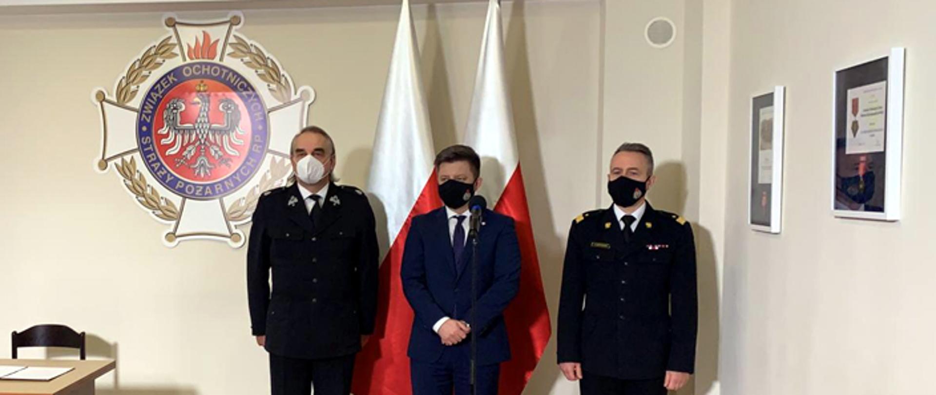 Na zdjęciu widnieje trzech mężczyzn . Od lewej ubrany w mundur wyjściowy Ochotniczej Straży Pożarnej prezes Zarządu Głównego druh Waldemar Pawlak. Kolejno po jego lewej stronie przed mikrofonem zabiera głos szef Kancelarii Prezesa Rady Ministrów Michał Dworczyk. Najbardziej po prawej na zdjęciu stoi w mundurze wyjściowym nadbrygadier Andrzej Bartkowiak. Wszyscy widoczni na zdjęciu mężczyźni noszą maseczki. Na ścianie po prawej stronie zawieszone dwa obrazy, na ścianie za mężczyznami wisi znak Związku Ochotniczych Straży Pożarnych. W lewej części widać część drewnianego biurka.