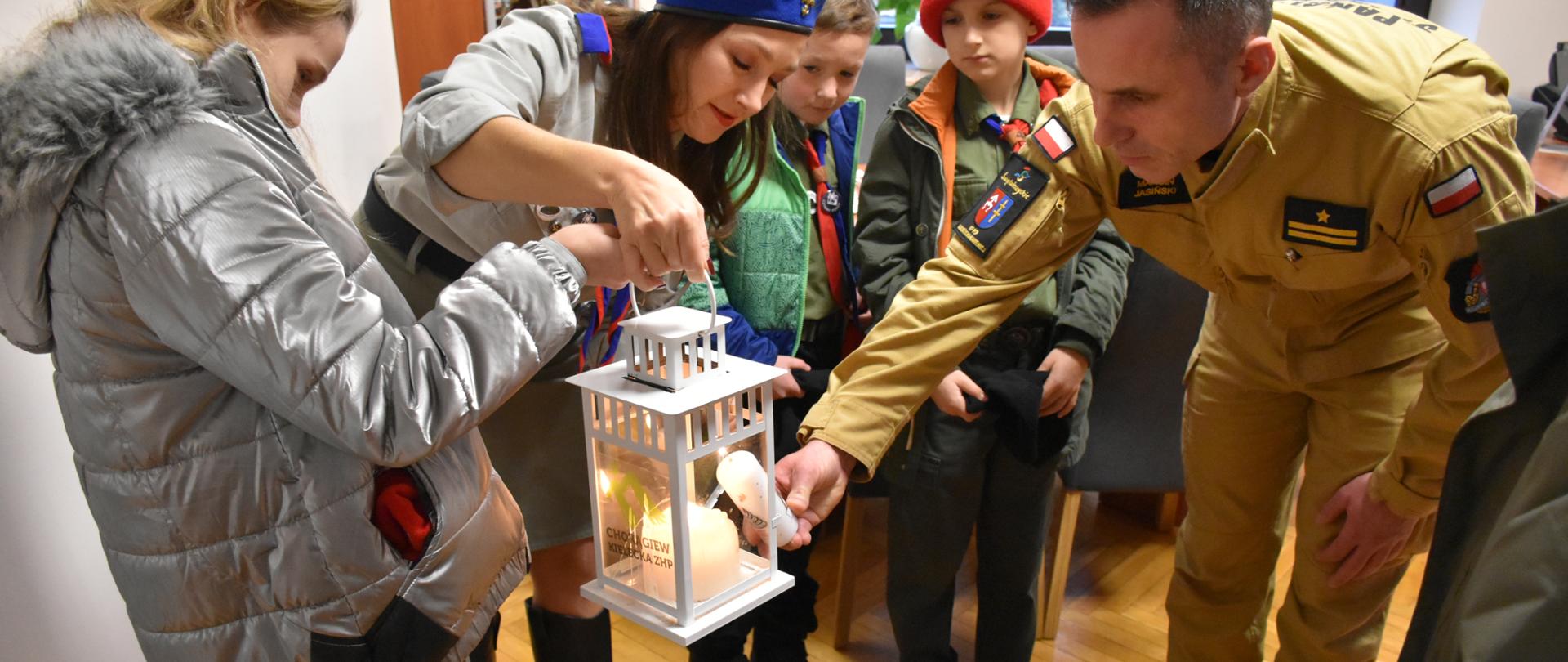 Zdjęcie obrazuje harcerzy ZHP Hufca Skarżysko-Kamienna, którzy przekazali Komendantowi Powiatowemu PSP w Skarżysku-Kamiennej Betlejemskie Światło Pokoju. Ze względu na trwającą pandemię wszyscy uczestnicy spotkania mają zasłonięte usta i nos maseczkami ochronnymi.