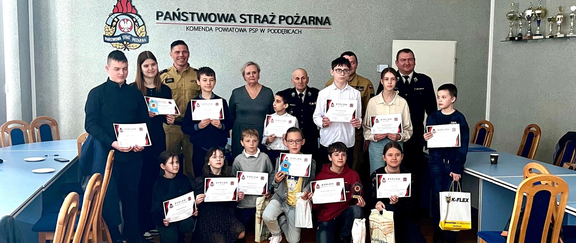 Zdjęcie grupowe dzieci i strażaków na sali w straży. 13 dzieci trzyma dyplomy kilku z nich kuca w pierwszym rzędzie reszta stoi