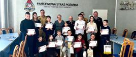 Zdjęcie grupowe dzieci i strażaków na sali w straży. 13 dzieci trzyma dyplomy kilku z nich kuca w pierwszym rzędzie reszta stoi