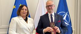 Kobieta w białym garniturze pozuje do zdjęcia z mężczyzną w garniturze, wojewodą, który trzyma odznaczenie 