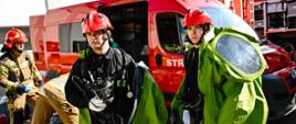 Dwóch strażaków ubranych w czarne ubrania koszarowe oraz zielone ubrania gazoszczelne idą ze sprzętem, na głowach mają czerwone kaski ubrania gazoszczelne ubrane są do połowy. W tle widać samochody pożarnicze oraz innych strażaków.