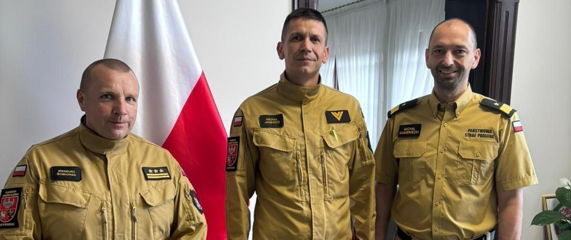 Trzech strażaków w jasnych mundurach. Od lewej Warmińsko Mazurski Komendant Wojewódzki nadbryg. Michał Kamieniecki, st. ogn. Michał Janewicz, i Komendant Powiatowy st. bryg Ireneusz Ścibiorek. W tle flaga Polski.