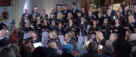Zdjęcie przedstawia chór śpiewający w Kościele i kilku członków orkiestry na pierwszym planie, członkowie mają w rękach instrumenty muzyczne.