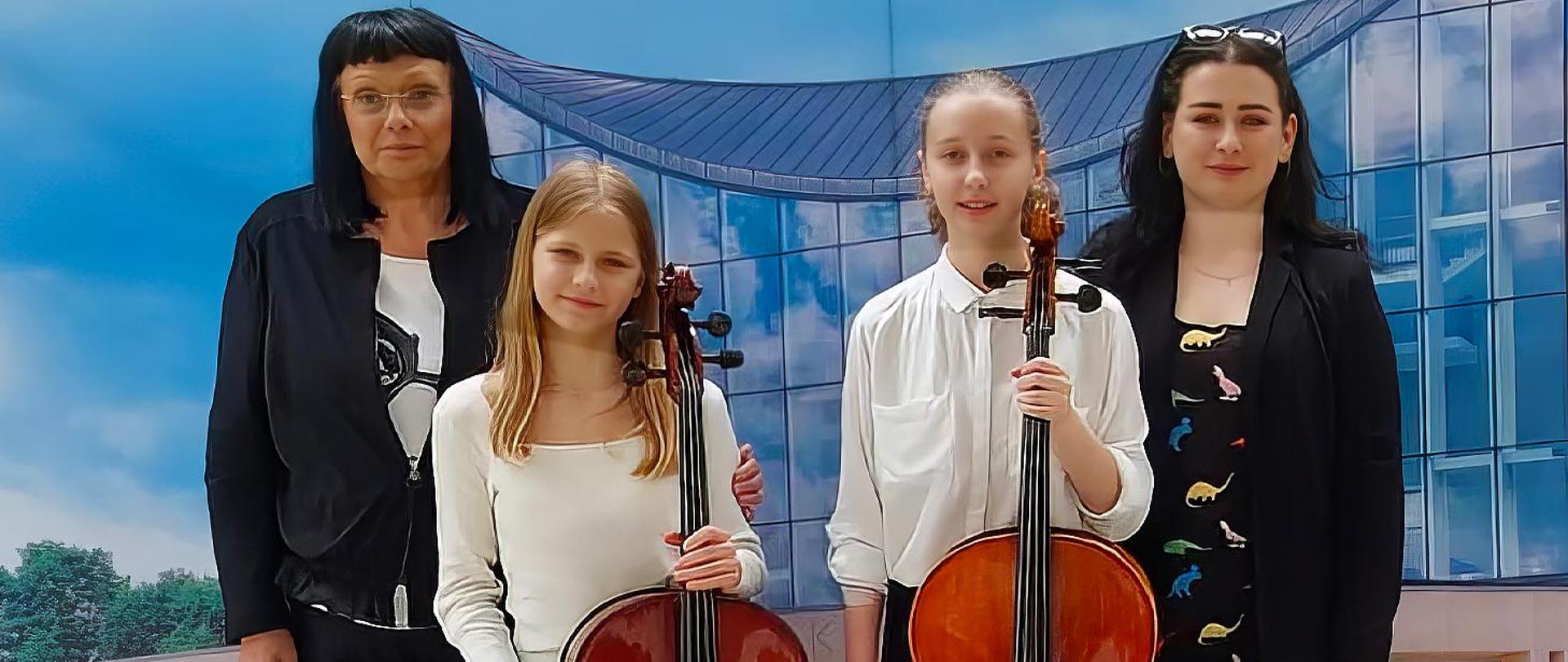 Na zdjęciu dwie dorosłe kobiety a pomiędzy nimi dwie dziewczyny trzymające w rękach wiolonczele.