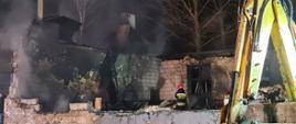 Zdjęcie przestawia funkcjonariuszy Państwowej Straży Pożarnej oraz Ochotniczej Straży Pożarnej podczas działań gaśniczych. W tle pogorzelisko remizy - zniszczony obiekt. 