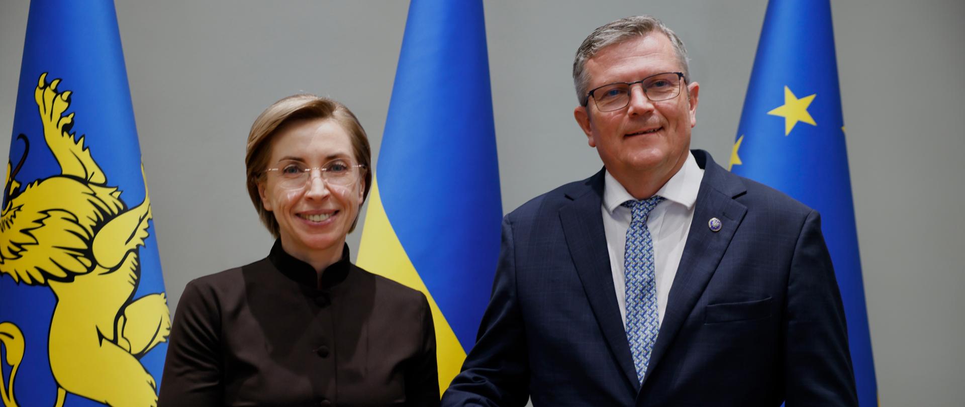 Na zdjęciu są dwie osoby - kobieta w brązowym stroju i mężczyzna w granatowym garniturze. Stoją obok siebie i patrzą w kierunku kamery. Za nimi są trzy flagi: Lwowa, Ukrainy i UE