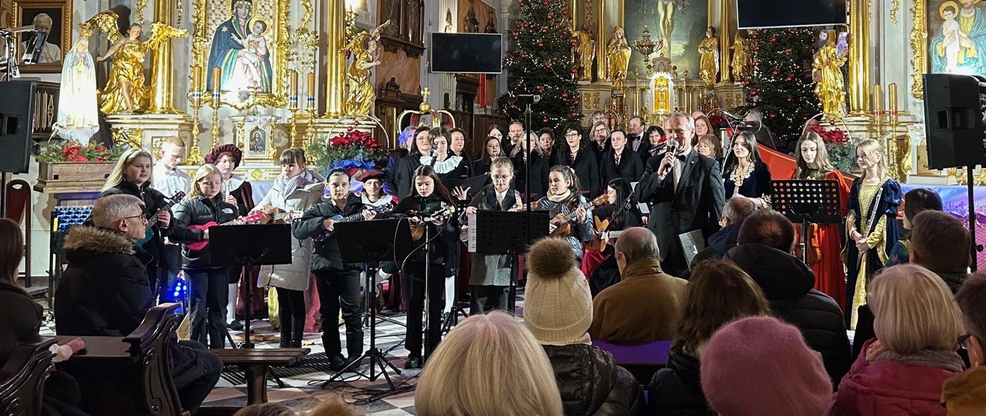 Wnętrze bazyliki wypełnione publicznością i wykonawcami podczas koncertu kolęd; na prezbiterium występują zespoły muzyki dawnej i chór.