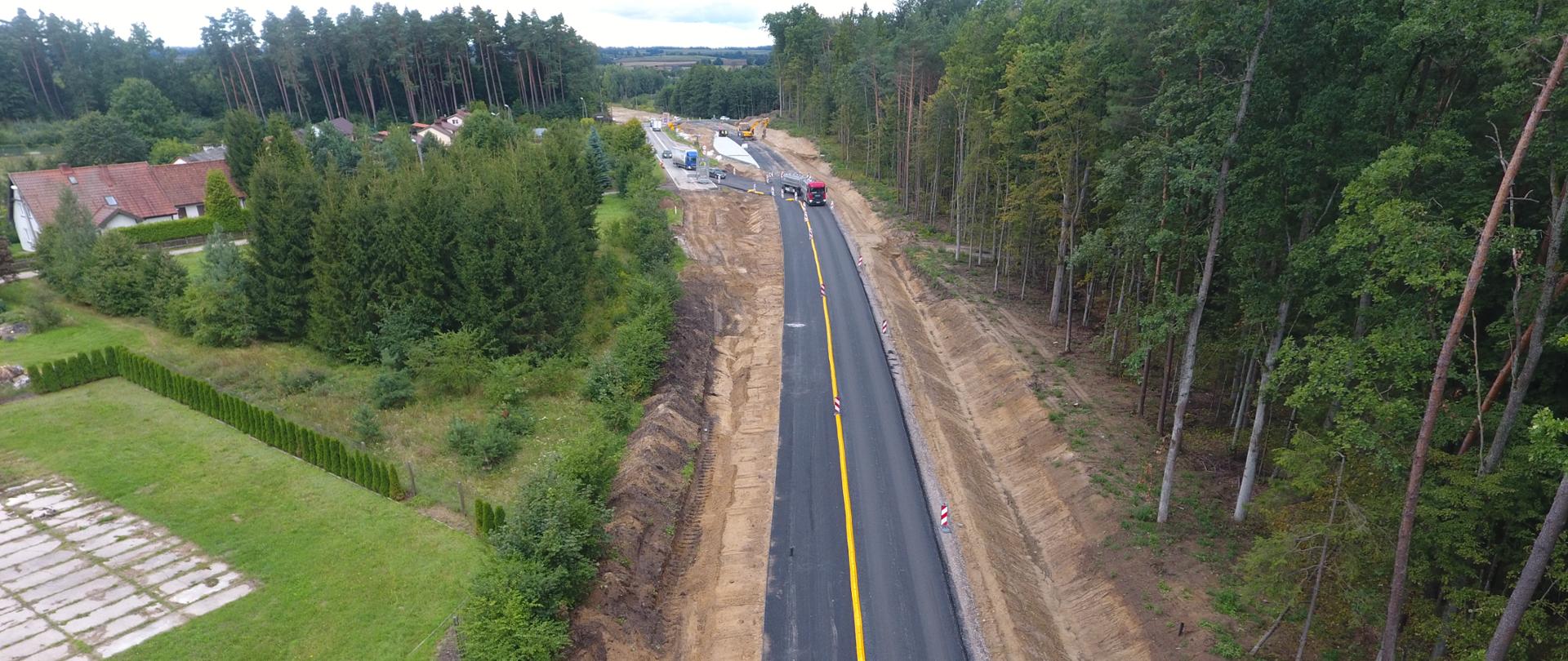 Na zdjęciu widoczna nitka drogi, z żółtym pasem wydzielającym kierunek ruchu pojazdów. Po prawej stronie widniej fragment lasu po lewej zabudowania ukryte za drzewostanem. Na drodze w oddali widać poruszające się pojazdy.
