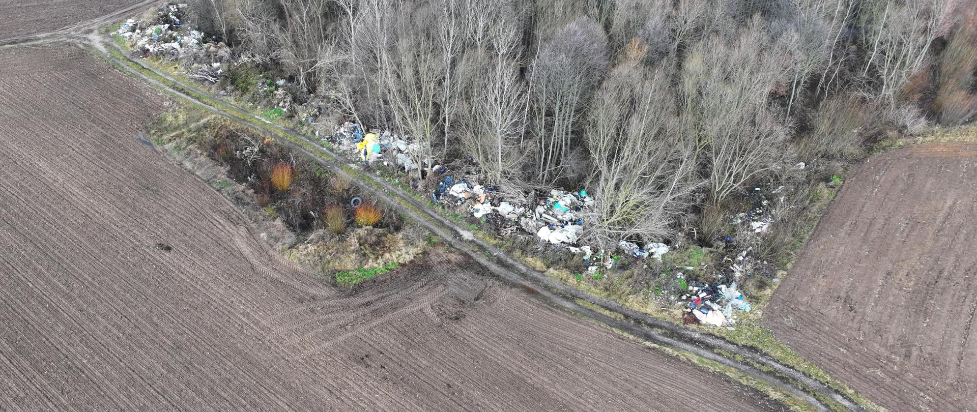 Widok z drona przedstawiający zaorane pola uprawne i skraj lasu z widocznymi nielegalnie zgromadzonymi odpadami zalegającymi wzdłuż polnej drogi.