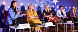 Na krzesłach ustawionych na podium siedzi pięć kobiet, z których jedna mówi do trzymanego w ręku mikrofonu