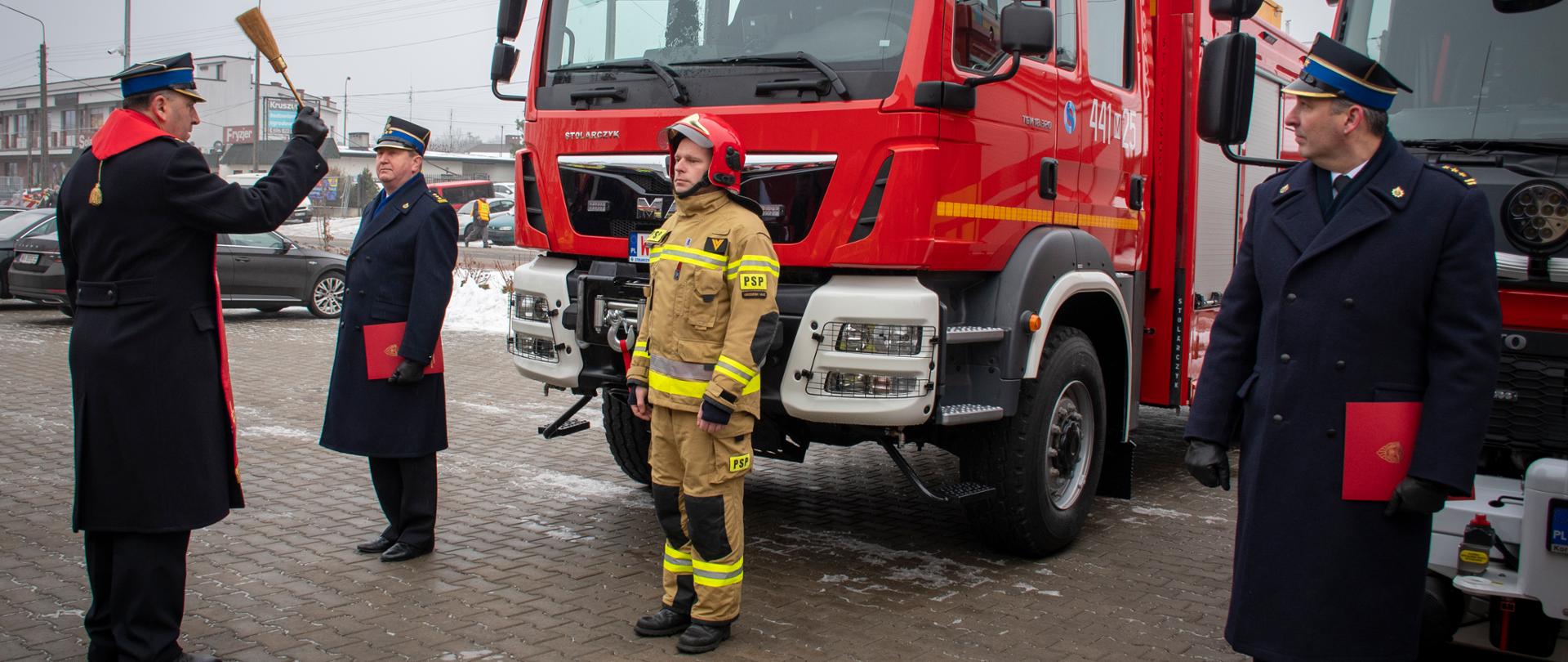 Poświęcenie przez kapelana straży pożarnej nowego ciężkiego samochodu ratowniczo-gaśniczego dla PSP w Grodzisku Mazowieckim. Przed samochodem stoi święcący go kapelan straży pożarnej, komendant powiatowy PSP w Grodzisku Mazowieckim, st. bryg Krzysztof Tryniszewski oraz kierowca z JRG w Grodzisku Mazowieckim.