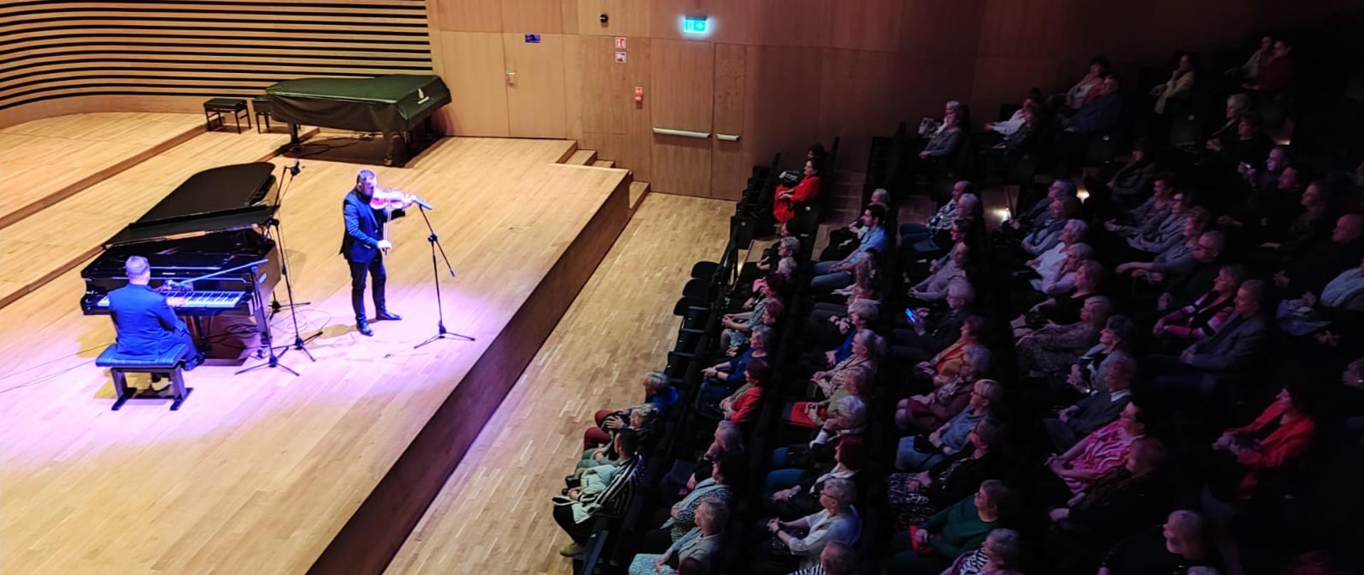 Widok z góry z lewej strony na scenę, na której mężczyzna gra na fortepianie, a drugi stojąc gra na skrzypcach. Po prawej stronie widać publiczność siedzącą na widowni.