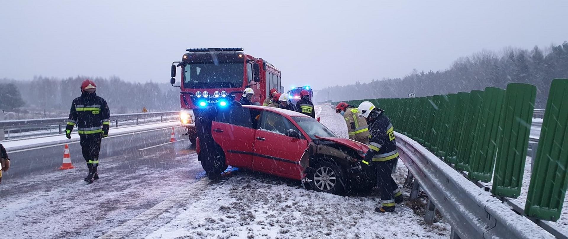Zdjęcie obrazuje uszkodzony przód samochodu osobowego marki seat Ibiza, który uderzył w barierę energochłonną rozdzielającą dwa kierunki jazdy. Przód samochodu został całkowicie zniszczony. W dalszym tle znajdują się pojazdy straży pożarnej. 