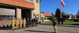 Strażacy stoją w szeregu i salutują w trakcie podnoszenia flagi państwowej na maszt