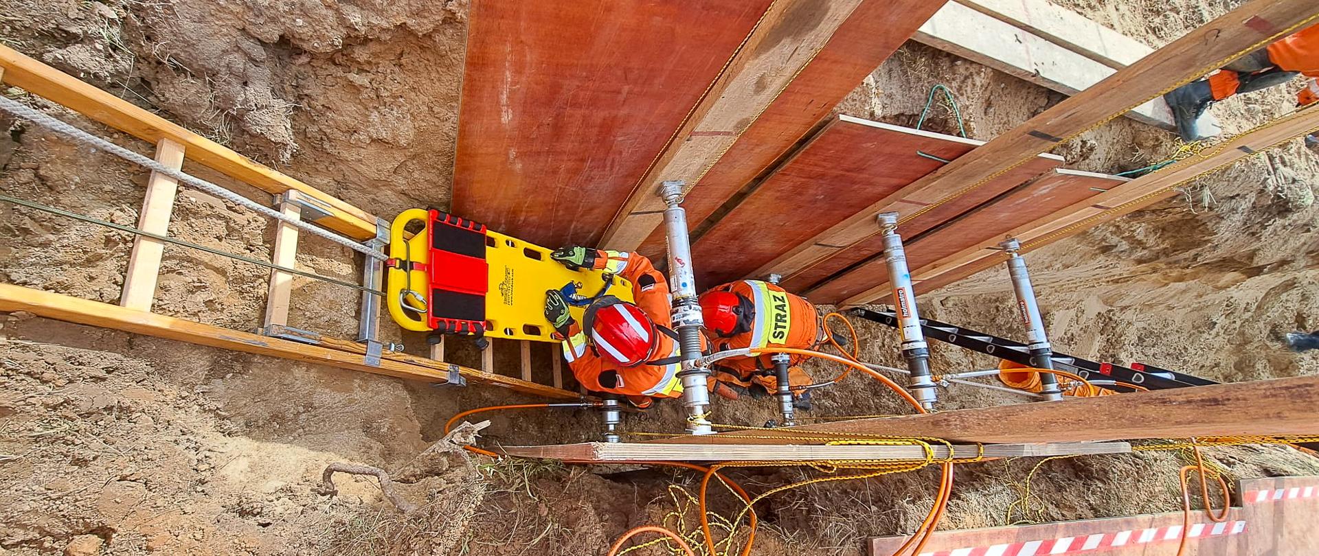 Dwóch ratowników w pomarańczowych kombinezonach i kaskach zabezpiecza i ewakuuje poszkodowanego z wykopu przy użyciu noszy i sprzętu stabilizacyjnego.