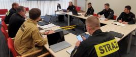 Na zdjęciu widać 7 strażaków państwowej straży pożarnej siedzących przy stole wraz z zastępcą dyrektora Biura Planowania Operacyjnego. Na stole monitory i laptopy. W tle prezentacja.