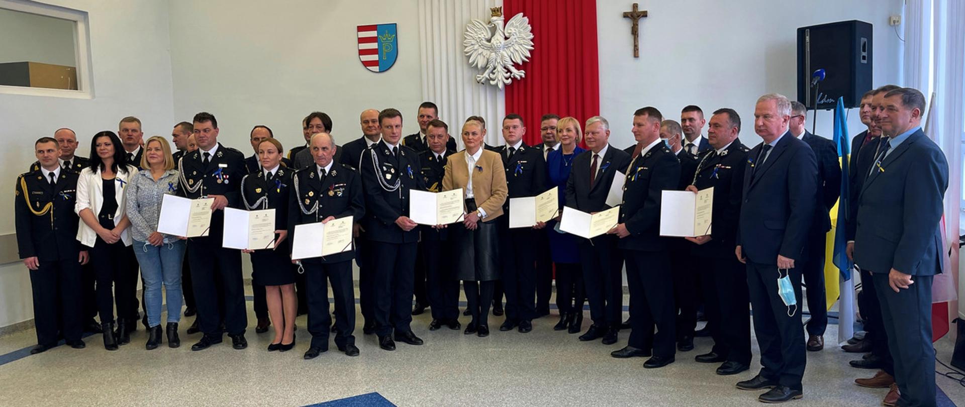 zdjęcie grupowe w pomieszczeniu Starostwa Powiatowego w Przysusze, na którym stoją druhny i druhowie OSP, Komendanci Powiatowi PSP, przedstawiciele samorządów terytorialnych jednostek którym wręczono promesy oraz Mazowiecki Komendant Wojewódzki i zaproszeni posłanki i posłowie RP