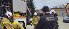 Zdjęcie przedstawia grupę strażaków zebranych wokół przenośnego, żółtego agregatu prądotwórczego marki Eisemann na betonowym placu manewrowym. Jeden ze strażaków pochyla się nad urządzeniem, podczas gdy pozostali – w tym mężczyzna na pierwszym planie w ciemnej kurtce z napisem „PAŃSTWOWA STRAŻ POŻARNA” oraz strażacy w piaskowych mundurach bojowych z oznaczeniem „OSP” – uważnie obserwują jego działanie. W tle widoczny jest bok otwartego wozu strażackiego, budynki jednostki oraz zaparkowany czarny samochód marki Audi pod pochmurnym niebem.