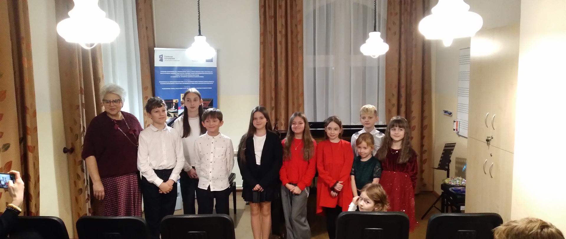 Zdjęcie przedstawia grupę około dziesięciorga dzieci w wieku szkolnym oraz starszą kobietę, stojących razem w kameralnej sali. Główny plan: Grupa osób stoi w rzędzie, uśmiechając się do aparatu. Są ubrani odświętnie: chłopcy w białych koszulach, dziewczynki w kolorowych sukienkach (czerwonej, bordowej, zielonej) lub ciemnych spódnicach. Starsza kobieta w bordowym swetrze i okularach stoi po lewej stronie grupy. Tło: Za plecami grupy znajduje się czarny fortepian oraz niebiesko-biały baner informacyjny z logotypami. Ściany są jasne, a w oknach wiszą brązowe zasłony. Z sufitu zwisają trzy białe, ozdobne lampy w kształcie talerzy. Pierwszy plan: W dolnej części kadru widoczne są tyły czarnych krzeseł ustawionych dla widowni oraz dłoń osoby fotografującej telefonem po lewej stronie. Całość sprawia wrażenie pamiątkowego zdjęcia wykonanego po koncercie lub uroczystości szkolnej w sali muzycznej.