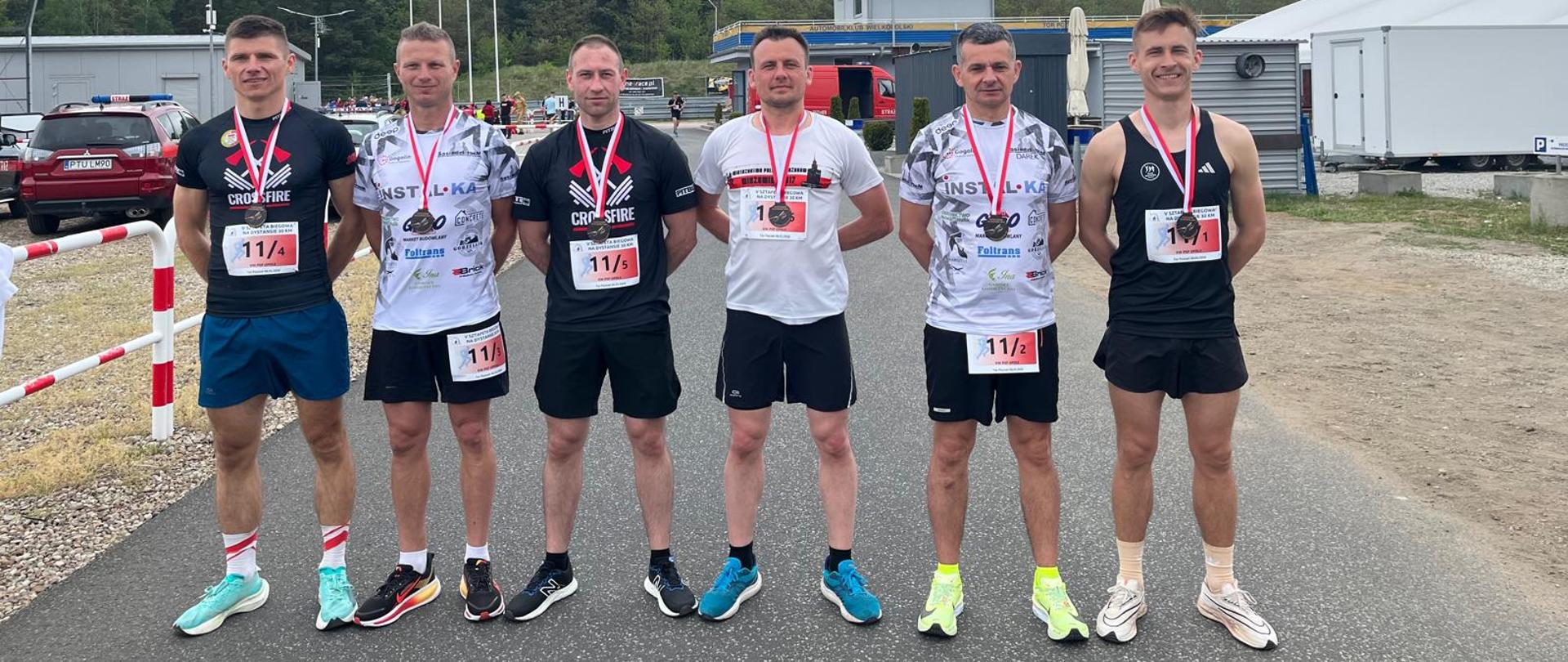 Sześciu strażaków z opolskiej reprezentacji stoi ramię w ramię na terenie toru wyścigowego w Przeźmierowie. Każdy ma na szyi medal i numer startowy. Za nimi znajduje się budynek z napisem „TOR POZNAŃ”, namioty organizatorów i pojazdy techniczne. Zdjęcie przedstawia drużynę po ukończeniu biegu sztafetowego.