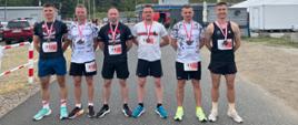 Sześciu strażaków z opolskiej reprezentacji stoi ramię w ramię na terenie toru wyścigowego w Przeźmierowie. Każdy ma na szyi medal i numer startowy. Za nimi znajduje się budynek z napisem „TOR POZNAŃ”, namioty organizatorów i pojazdy techniczne. Zdjęcie przedstawia drużynę po ukończeniu biegu sztafetowego.
