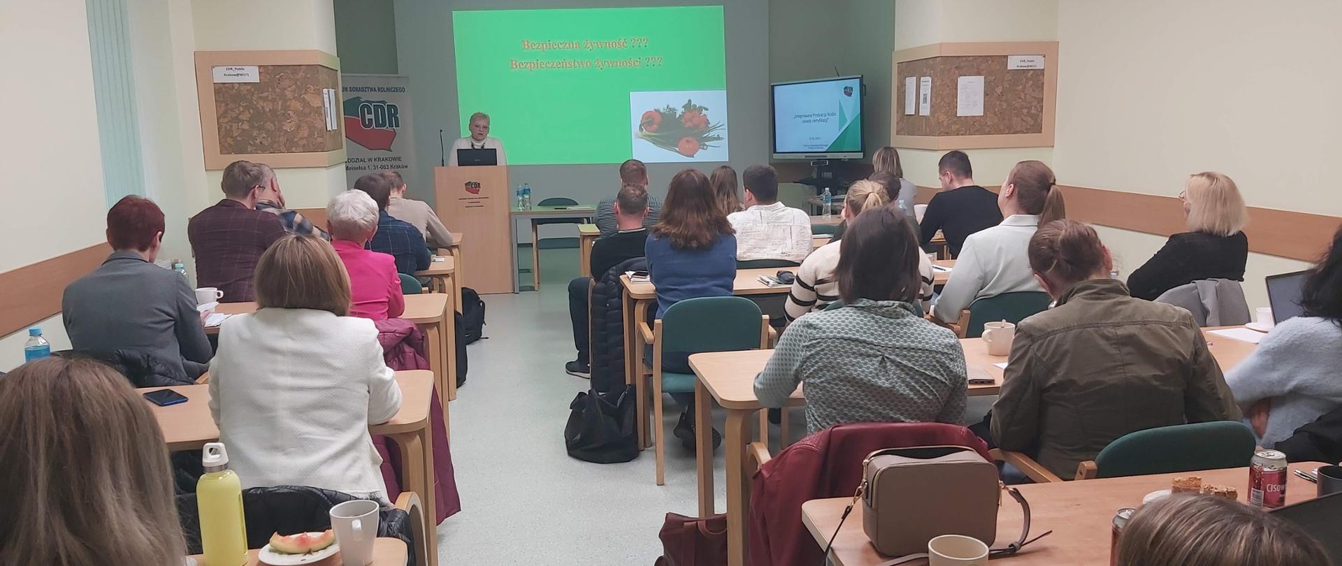 Na zdjęciu widoczna kobieta prowadząca szkolenie na tle ekranu z wyświetloną prezentacją a także sala wykładowa z widoczną grupą uczestników szkolenia siedzącą przy stolikach, zwróconymi w kierunku prowadzącej 